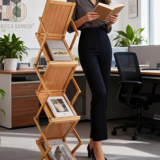 Porte-revues en bois et bambou. Un présentoir pliable en accordéon avec plusieurs étagères inclinées contenant des magazines, posé au sol à côté d'une femme lisant dans un bureau.