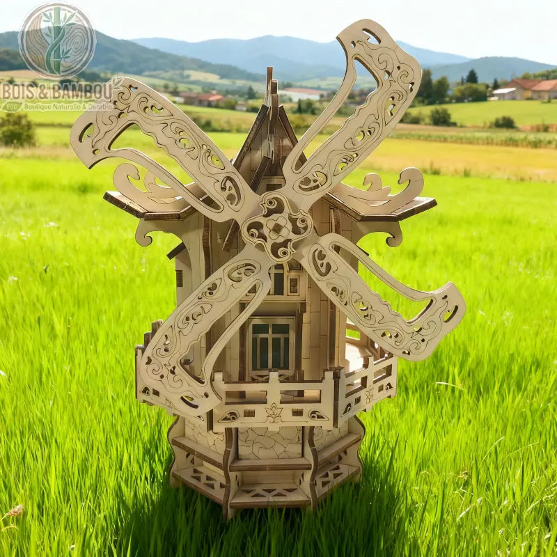 Puzzle 3D moulin à vent en bois. Maquette détaillée en bois découpé au laser avec pales sculptées, posée dans un champ d'herbe verte sous un ciel bleu et des montagnes.