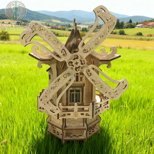 Puzzle 3D moulin à vent en bois. Maquette détaillée en bois découpé au laser avec pales sculptées, posée dans un champ d'herbe verte sous un ciel bleu et des montagnes.