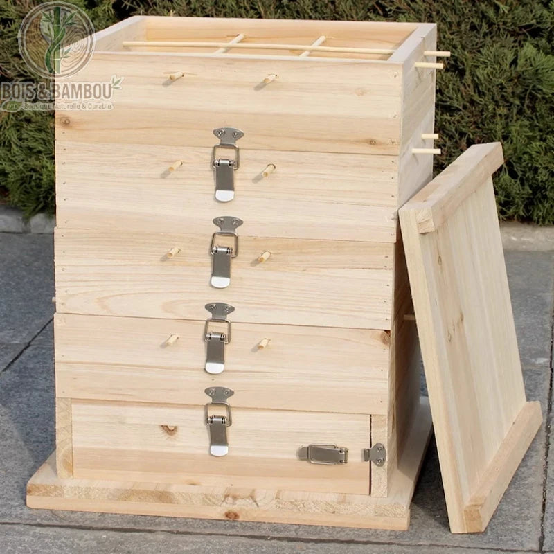 Ruche Traditionnelle en Cèdre. Ruche à plusieurs étages en bois clair avec fermoirs métalliques et chevilles en bois, posée sur un sol en pierre devant des arbustes.