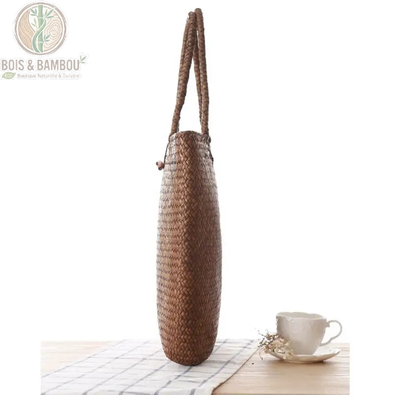 Sac a main en paille tressee lateral. Vue de profil d'un sac cabas en paille tressée marron foncé, posé sur une nappe à carreaux à côté d'une tasse blanche, illustrant sa finesse et sa souplesse.