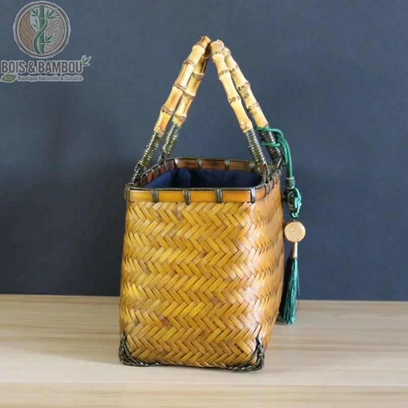 Sac cabas Coffret en bambou claire lateral. Vue de profil d'un sac rectangulaire en bambou tressé naturel avec double anse rigide, doublure intérieure bleue et pompon vert décoratif sur fond sombre.
