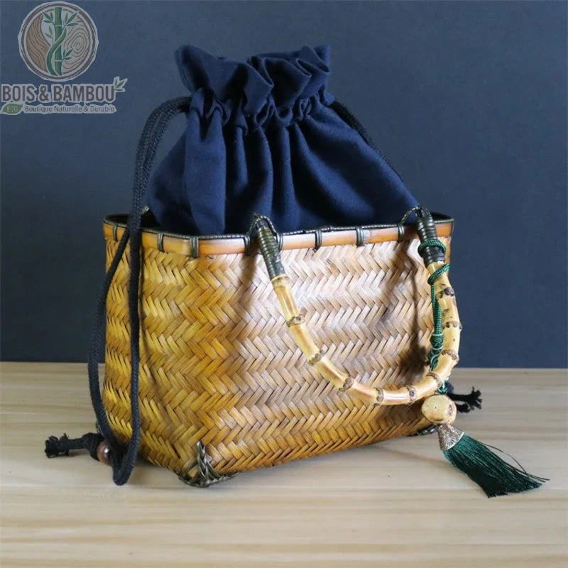Sac cabas Coffret en bambou claire. Sac artisanal rectangulaire en bambou tressé avec poignée, pochon bleu marine et pompon, posé sur une table en bois sur un fond sombre.