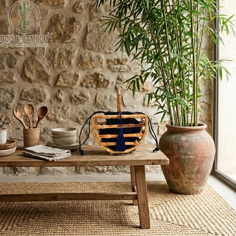 Sac corbeille en bambou a pompon. Sac artisanal en tiges de bambou avec poignée et pochon noir à pompon, posé sur une table en bois rustique dans un décor naturel avec mur en pierre et plante.