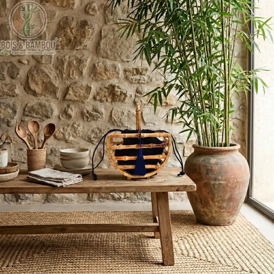 Sac corbeille en bambou a pompon. Sac artisanal en tiges de bambou avec poignée et pochon noir à pompon, posé sur une table en bois rustique dans un décor naturel avec mur en pierre et plante.