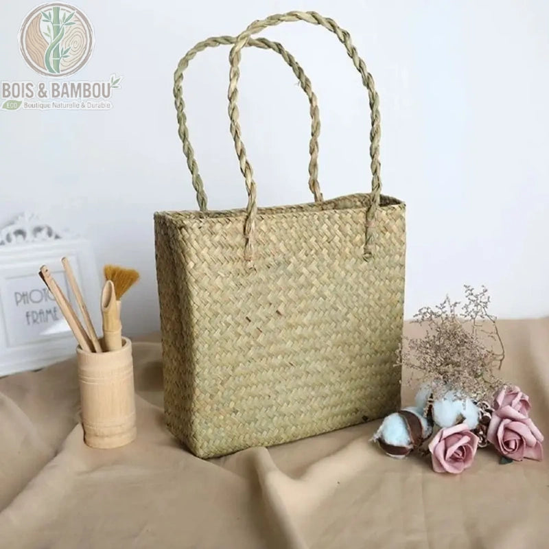 Sac fourre tout en paille et bambou couleur claire. Un sac cabas rectangulaire en paille tressée de couleur beige clair avec anses souples, présenté à côté d'un pot en bambou et de fleurs séchées.