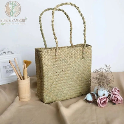 Sac fourre tout en paille et bambou couleur claire. Un sac cabas rectangulaire en paille tressée de couleur beige clair avec anses souples, présenté à côté d'un pot en bambou et de fleurs séchées.