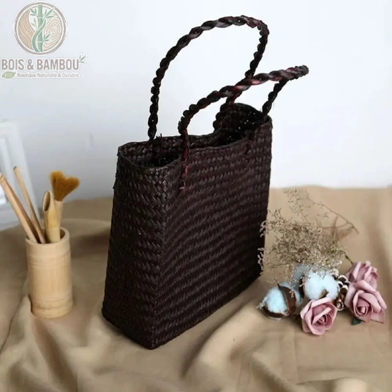 Sac fourre tout en paille et bambou couleur marron. Sac cabas rectangulaire en paille tressée marron foncé avec anses souples, posé sur un tissu beige à côté d'accessoires en bambou et de fleurs.