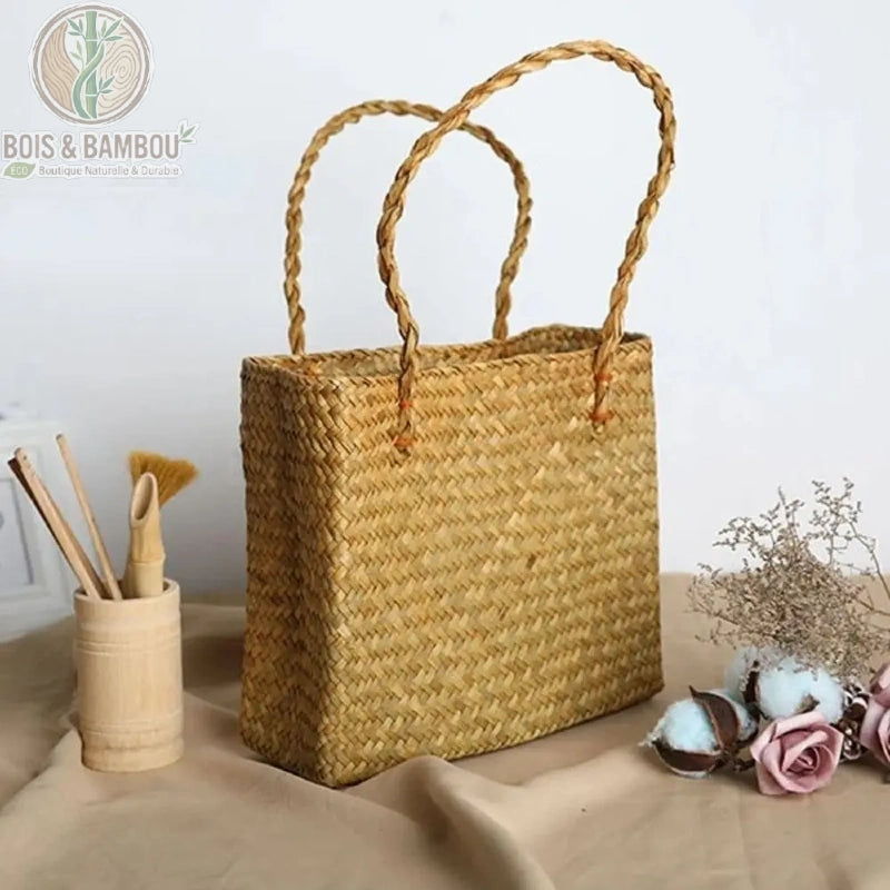 Sac fourre tout en paille et bambou couleur paille. Un sac cabas rectangulaire en paille tressée naturelle avec anses souples, présenté à côté d'un pot en bambou et de fleurs de coton décoratives.