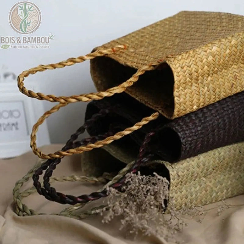 Sac fourre tout en paille et bambou situation 1. Plusieurs sacs cabas artisanaux en paille tressée aux teintes naturelles et chocolat empilés à plat, mettant en avant leurs anses tressées souples.