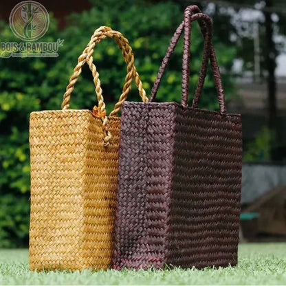 Sac fourre tout en paille et bambou situation 2. Deux sacs cabas artisanaux rectangulaires en paille tressée, l'un naturel et l'un marron foncé, posés côte à côte sur une pelouse verte.