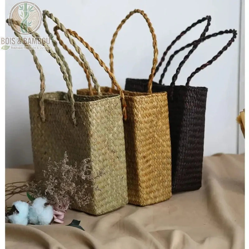 Sac fourre tout en paille et bambou. Alignement de quatre sacs cabas artisanaux en paille tressée déclinés en nuances de beige et marron, présentés avec des fleurs de coton sur un drapé neutre.
