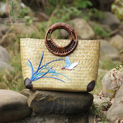 Sac main retro en bambou tisse situation 1. Un sac à main artisanal en bambou tressé avec anses rondes sombres et broderies d'oiseau et fleurs bleues, posé sur des rochers dans un cadre naturel.