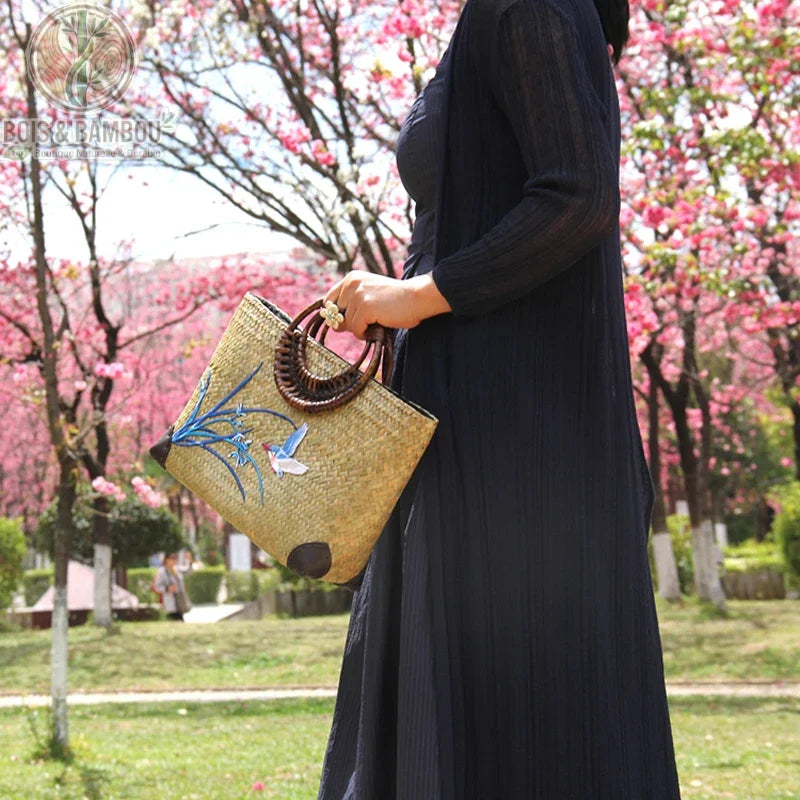 Sac main retro en bambou tisse situation 2. Femme vêtue d'une robe noire portant un sac artisanal en bambou avec broderies d'oiseau et fleurs, devant des cerisiers en fleurs dans un parc ensoleillé.