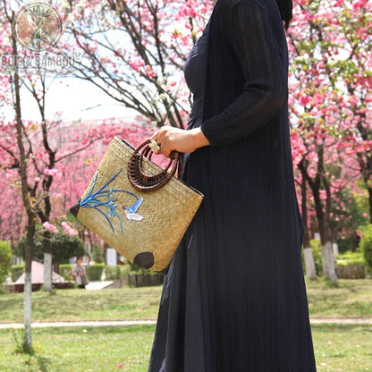 Sac main retro en bambou tisse situation 2. Femme vêtue d'une robe noire portant un sac artisanal en bambou avec broderies d'oiseau et fleurs, devant des cerisiers en fleurs dans un parc ensoleillé.