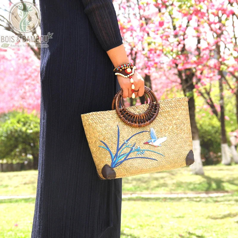 Sac main retro en bambou tisse situation 3. Femme portant un sac cabas artisanal en bambou tressé, orné de broderies d'un martin-pêcheur et de fleurs bleues, avec anses rondes en bambou sombre.