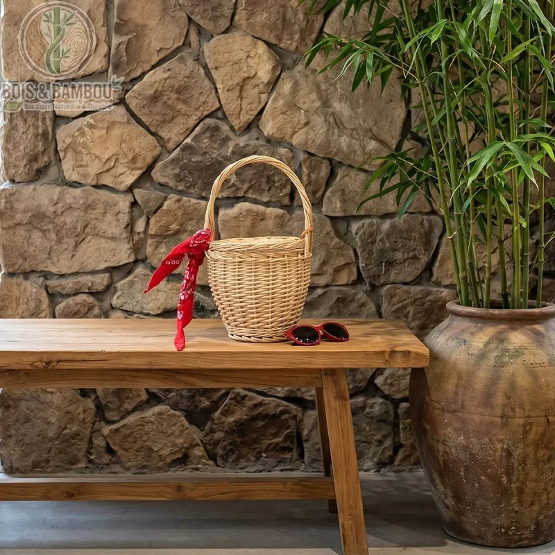 Sac panier en bambou et paille claire situation. Un panier seau en osier clair décoré d'un bandana rouge, posé sur un banc en bois rustique à côté de lunettes de soleil, devant un mur en pierres.