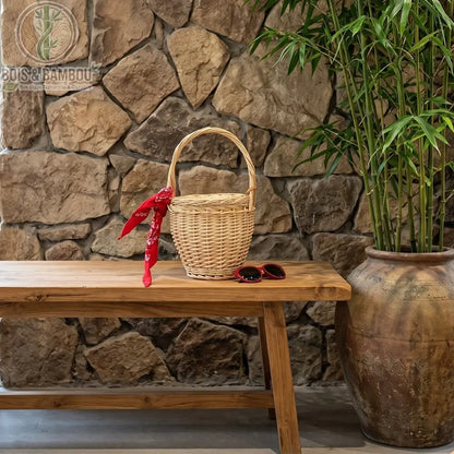 Sac panier en bambou et paille claire situation. Un panier seau en osier clair décoré d'un bandana rouge, posé sur un banc en bois rustique à côté de lunettes de soleil, devant un mur en pierres.