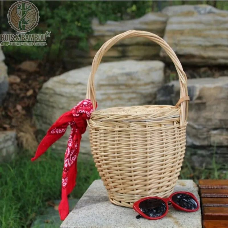 Sac panier en bambou et paille claire. Panier seau artisanal en osier clair avec couvercle, orné d'un bandana rouge et accompagné de lunettes de soleil rouges, posé sur un bloc de pierre en extérieur.