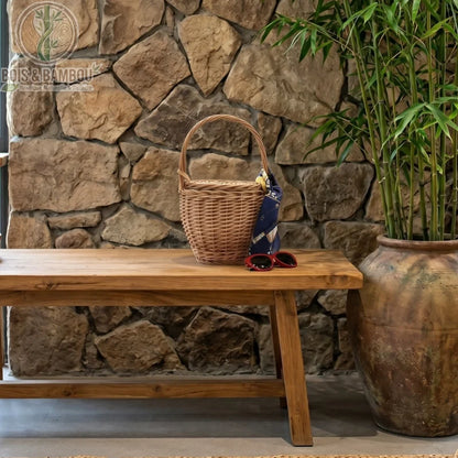 Sac panier en bambou et paille marron situation. Panier rond en osier tressé posé sur un banc en bois rustique devant un mur de pierres, décoré d'un foulard bleu et accompagné de lunettes de soleil.