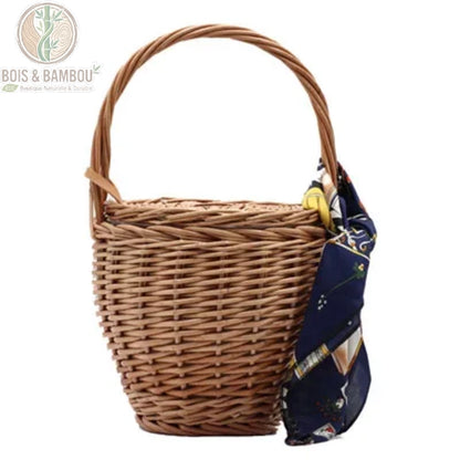 Sac panier en bambou et paille claire. Panier seau artisanal en osier clair avec couvercle, orné d'un bandana rouge et accompagné de lunettes de soleil rouges, posé sur un bloc de pierre en extérieur.