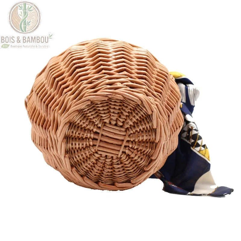 Sac panier en bambou et paille vue 1. Vue de dessous d'un panier seau en osier tressé marron montrant la base circulaire et le tressage artisanal serré, avec un pan de foulard bleu visible sur le côté.