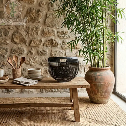 Sac panier en bambou tresse noir. Sac à main demi-lune en bambou noir brillant exposé sur un banc en bois rustique, dans un intérieur naturel avec mur en pierre et plante de bambou en pot.