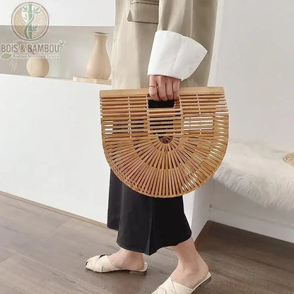 Sac panier en bambou tresse situation 2. Une personne en tenue élégante avec veste beige et robe noire tient un sac à main demi-lune en bambou ajouré, posé à plat sur un sol en bois clair.