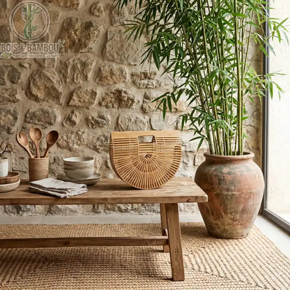 Sac panier en bambou tresse. Sac à main demi-lune en bambou ajouré posé sur un banc en bois rustique, dans un décor naturel avec mur en pierre, pot en terre cuite et plante de bambou.
