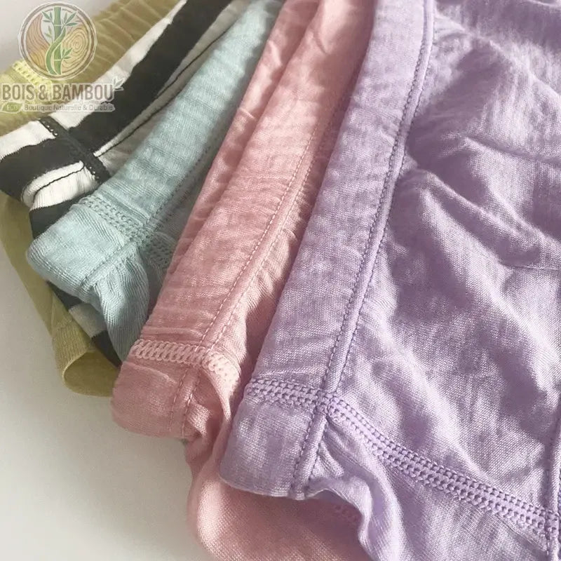 Slip bikini en laine mérinos et fibre de bambou. Gros plan sur une pile de sous-vêtements aux couleurs pastel (mauve, rose, bleu, rayé noir et blanc, vert) montrant la texture douce.