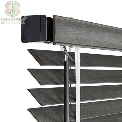 Vue détaillée du mécanisme supérieur de stores vénitiens en bois de tilleul gris foncé sur mesure. Boîtier de commande noir, tige et corde de levage. Lamelles texturées avec grain de bois.