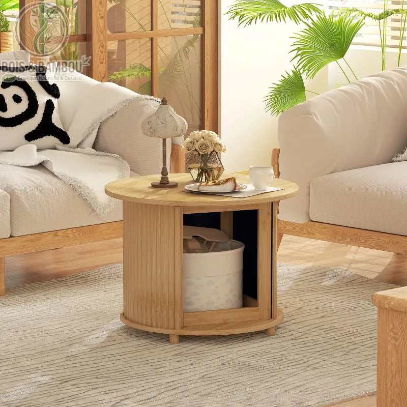 Table Basse en Bambou. Une table ronde en bois clair avec un compartiment de rangement ouvert, décorée d'une lampe et de fleurs, placée entre deux canapés dans un salon lumineux.