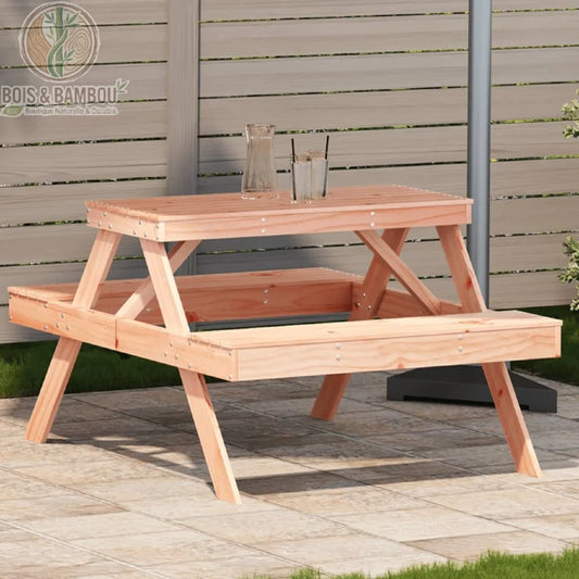 Table de Pique-nique en Douglas. Une table de jardin classique en bois clair avec bancs intégrés, disposée sur une terrasse en dalles de pierre devant une clôture en bois gris.