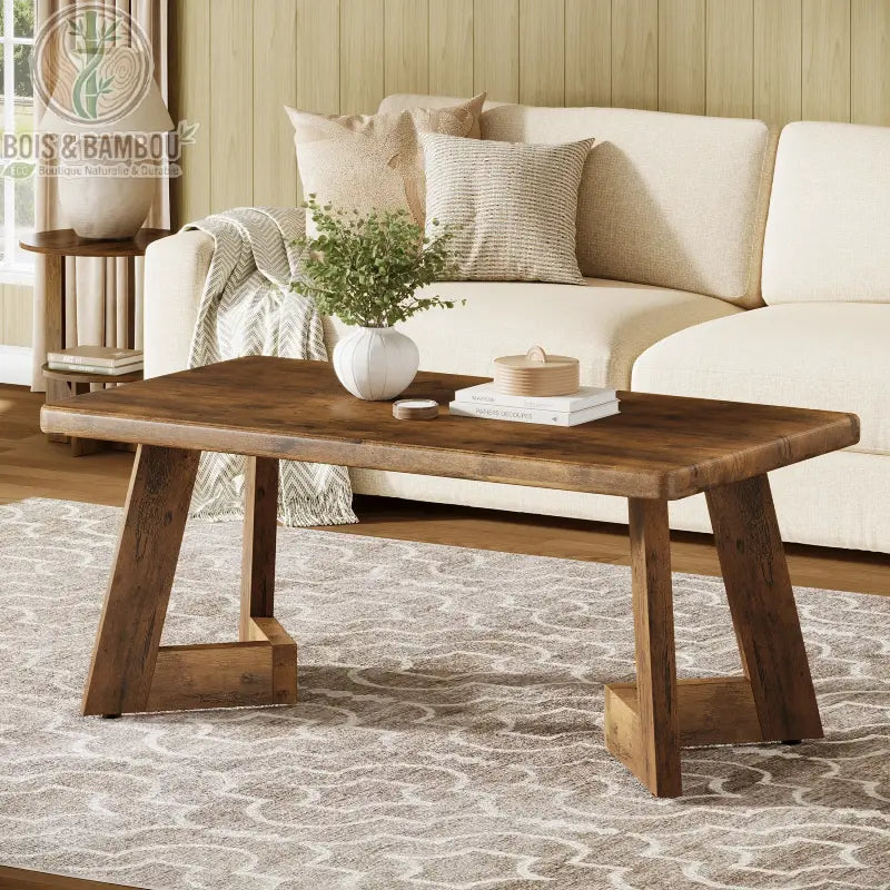 Table en bois massif basse style farmhouse 2. Table de salon rustique au fini bois chaleureux avec piètement angulaire distinctif, disposée sur un tapis à motifs devant un canapé crème.