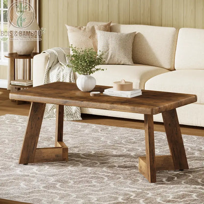 Table en bois massif basse style farmhouse 2. Table de salon rustique au fini bois chaleureux avec piètement angulaire distinctif, disposée sur un tapis à motifs devant un canapé crème.