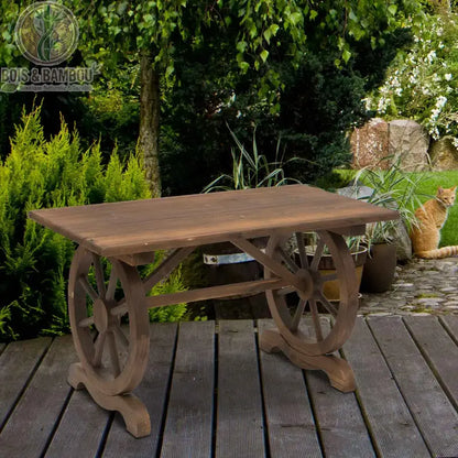 Table de Jardin Rustique. Une table en bois massif de style campagnard avec des pieds originaux en forme de roues de charrette, posée sur une terrasse en bois dans un jardin arboré.