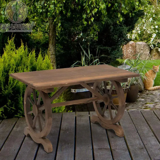 Table de Jardin Rustique. Une table en bois massif de style campagnard avec des pieds originaux en forme de roues de charrette, posée sur une terrasse en bois dans un jardin arboré.
