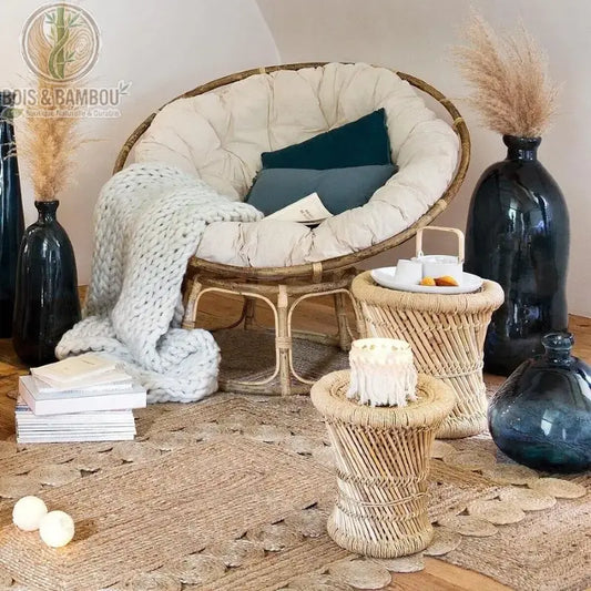 Tables Gigognes Ilyan. Deux tables d'appoint en rotin tressé de forme sablier, accompagnées d'un fauteuil papasan et de décoration bohème sur un grand tapis en jute.