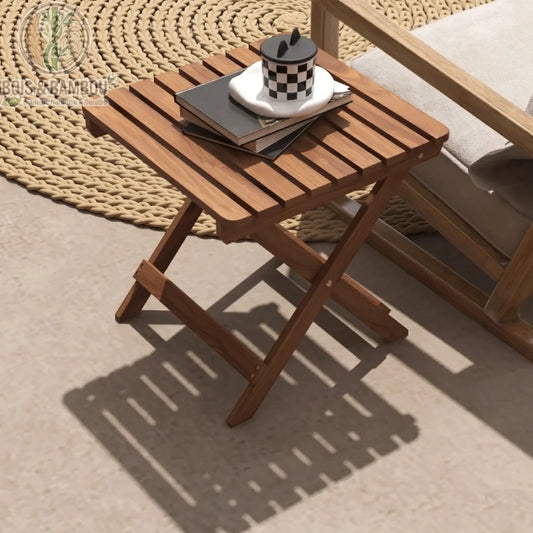 Tables de Patio en Acacia. Une petite table pliante carrée en lattes de bois brun, décorée de livres et d'une tasse, projetant son ombre sur une terrasse ensoleillée.