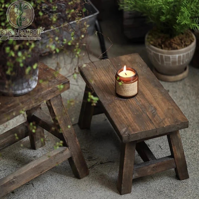 Tabouret bas en bois situation 2. Tabouret bas artisanal en bois foncé vieilli utilisé comme support décoratif pour une bougie parfumée allumée, créant une ambiance zen et chaleureuse.