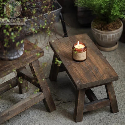 Tabouret bas en bois situation 2. Tabouret bas artisanal en bois foncé vieilli utilisé comme support décoratif pour une bougie parfumée allumée, créant une ambiance zen et chaleureuse.