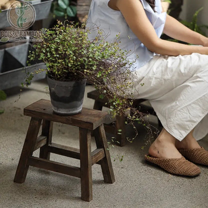 Tabouret bas en bois situation. Petit banc rustique en bois foncé supportant une plante grimpante en pot, avec une personne assise sur un second tabouret en arrière-plan dans une ambiance jardin.