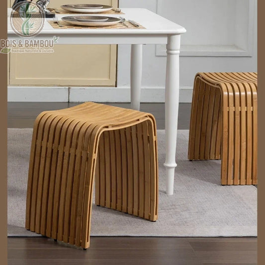 Tabouret moderne en bambou Nan. Deux tabourets au design incurvé et ajouré, composés de lattes de bois clair, disposés sur un tapis gris devant une table à manger blanche.