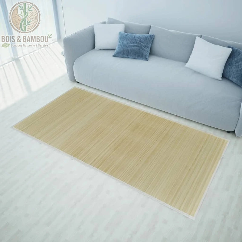 Tapis en bambou naturel de dimensions 100x160 cm, posé sur un sol en bois clair dans un salon moderne. Le tapis présente une texture à lattes horizontales claires avec une bordure en tissu beige fin, placé devant un canapé gris clair orné de coussins bleus et blancs.