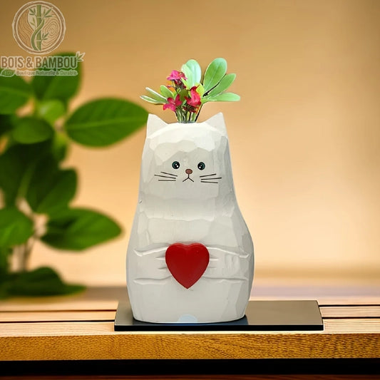Vase Statue de Chat en Bois Massif. Une figurine de chat blanche sculptée tenant un cœur rouge, servant de vase pour de petites fleurs, posée sur un socle noir devant un fond flou.