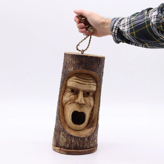 Nichoir en bois sculpté tenu à la main par sa corde de suspension, visage grincheux sculpté