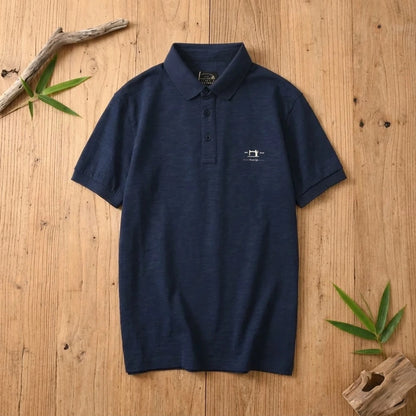 Polo homme bleu marine en coton et fibre de bambou posé à plat sur une table en bois brut avec feuilles de bambou