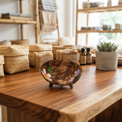 Porte-savon en noix de coco rond à fleurs peintes et fond percé, sur un comptoir en bois près de savons artisanaux