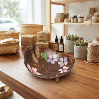 Porte-savon en noix de coco à bord découpé, fleurs mauves et rose peintes, posé sur petits pieds en bois