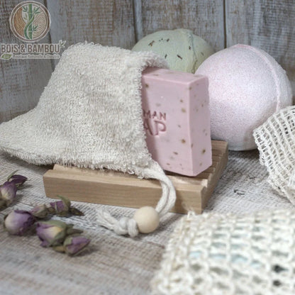 Sac à savon en tissu éponge écru posé sur un porte-savon en bois avec savon rose et boules de bain
